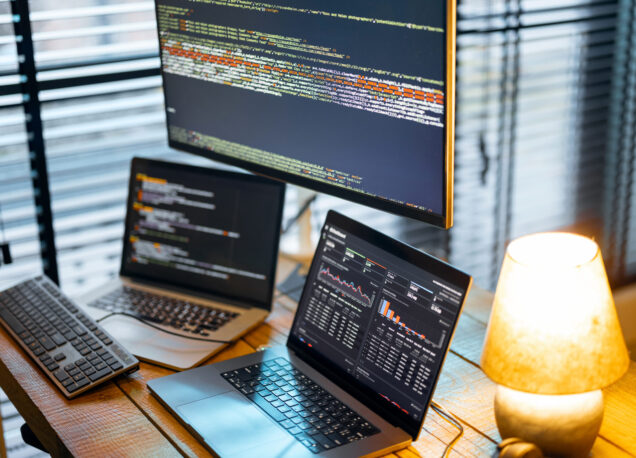 Cozy workplace with lapto and desktop computers with programming code on the screens. Programmer work table, Close-up view with no people